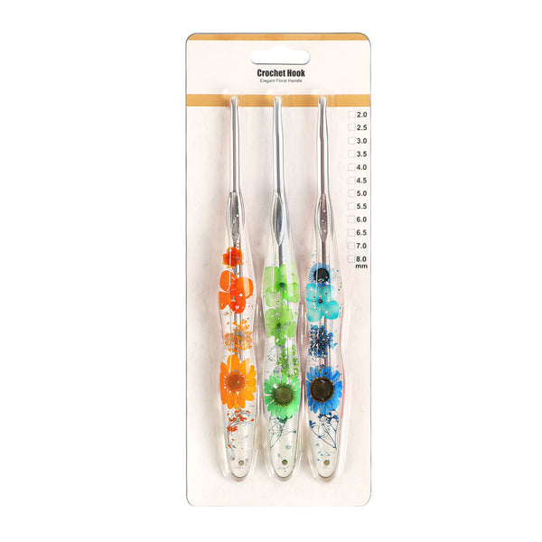 Three crochet hooks in a clear plastic case with printed size markings, hanging from a cardboard hanger. The handles are decorated with dried flowers in orange, green, and blue, with black metal tips.