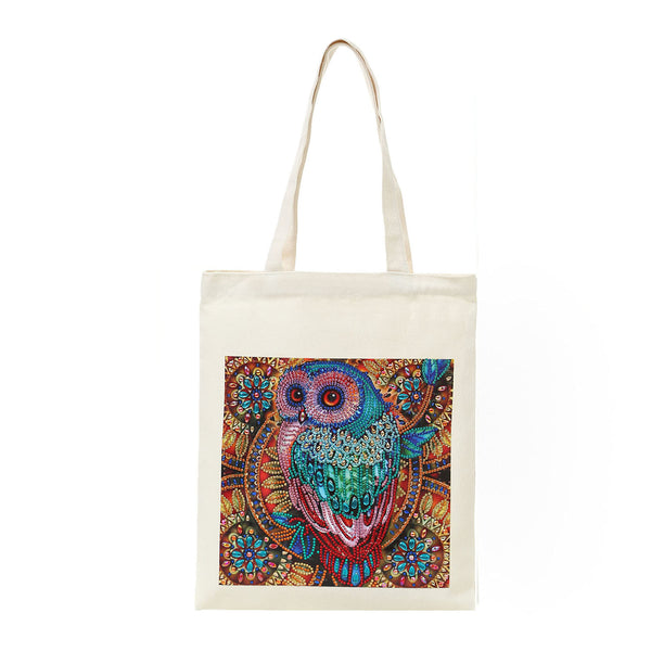 An off-white canvas tote bag with long handles, featuring a colorful, highly detailed owl illustration printed on the front. The owl is decorated with intricate patterns in shades of teal, blue, pink, and gold against a dark background.