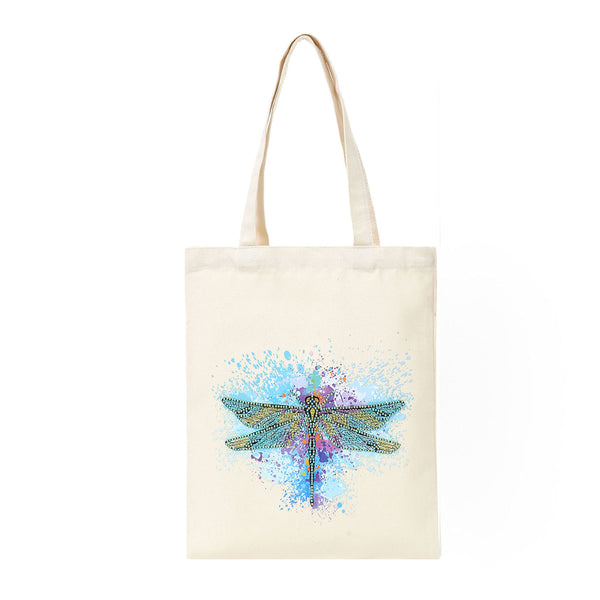 An off-white canvas tote bag with two shoulder straps, featuring a colorful watercolor-style dragonfly design in blue, purple, and green on the front, set against a plain white background.