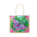 A square canvas tote bag with cream-colored rope handles, featuring a colorful beaded butterfly and leaf design on a pink glittery background.