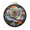 A circular embroidery hoop containing a detailed floral design featuring a large succulent flower in muted green, pink, and orange tones on a dark navy fabric background.