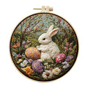 A circular embroidery piece held in a wooden hoop, featuring a white rabbit sitting among colorful flowers and patterned Easter eggs in a garden scene.