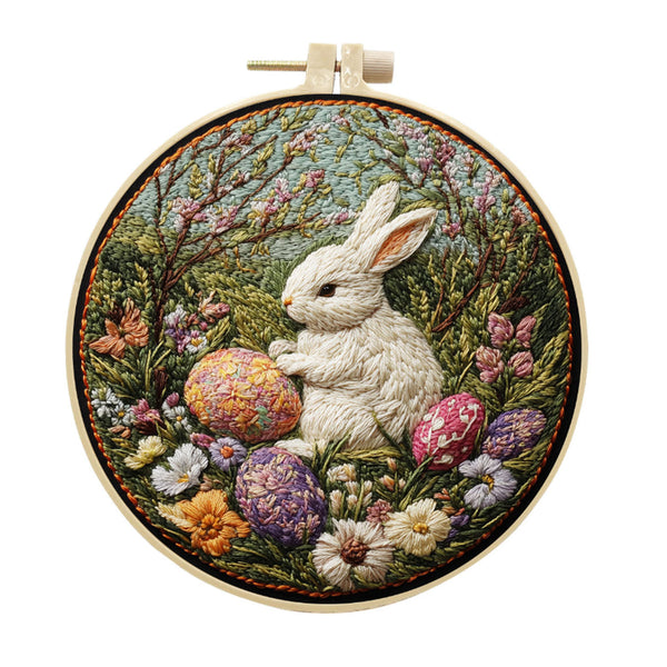 A circular embroidery piece held in a wooden hoop, featuring a white rabbit sitting among colorful flowers and patterned Easter eggs in a garden scene.