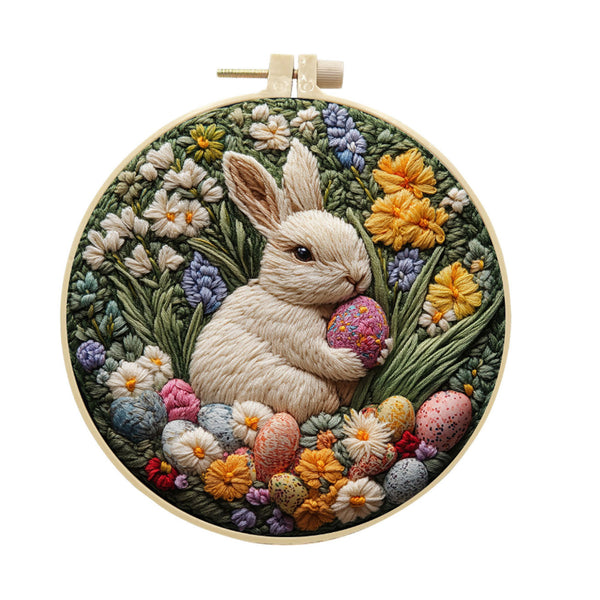 A circular embroidered wall hanging in a wooden hoop, featuring a cream-colored bunny holding a pink egg surrounded by colorful wildflowers and Easter eggs on a white background.