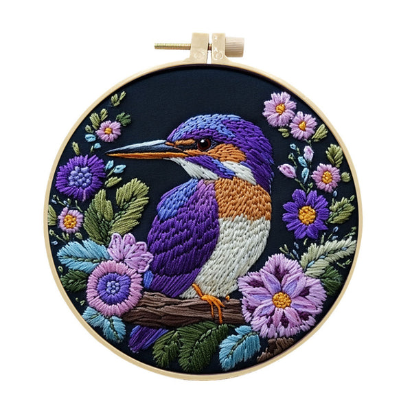 A circular embroidery hoop containing a colorful embroidered kingfisher surrounded by purple flowers and green leaves on a dark background, shown against a plain white backdrop.