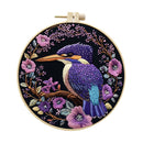 A circular embroidery hoop containing a detailed bird and floral design. The artwork features a purple and blue kingfisher perched on a branch, surrounded by pink and purple flowers on a black background, with a light wood hoop frame.