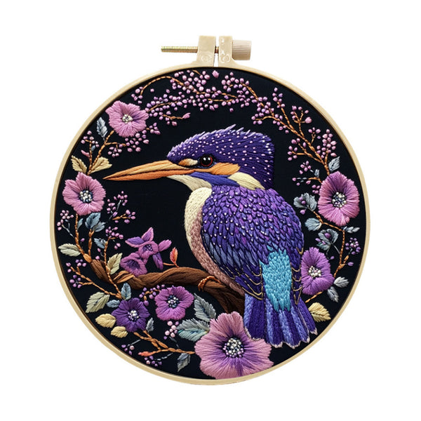A circular embroidery hoop containing a detailed bird and floral design. The artwork features a purple and blue kingfisher perched on a branch, surrounded by pink and purple flowers on a black background, with a light wood hoop frame.