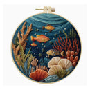 A circular embroidery hoop containing a detailed multicolor sea life embroidery design with orange and yellow fish, red and green coral, and beige seashells on a dark blue background.