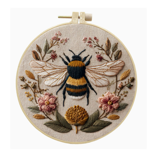 A circular embroidery hoop containing a detailed floral and insect textile design, featuring a bumblebee at the center surrounded by pink flowers, green leaves, and gold-toned botanical elements on a light beige fabric background.
