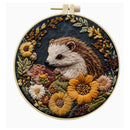 A circular embroidery hoop containing a detailed floral and hedgehog textile artwork. The design features a brown and cream hedgehog surrounded by yellow, pink, and white flowers with green and gold leaves on a dark navy background.