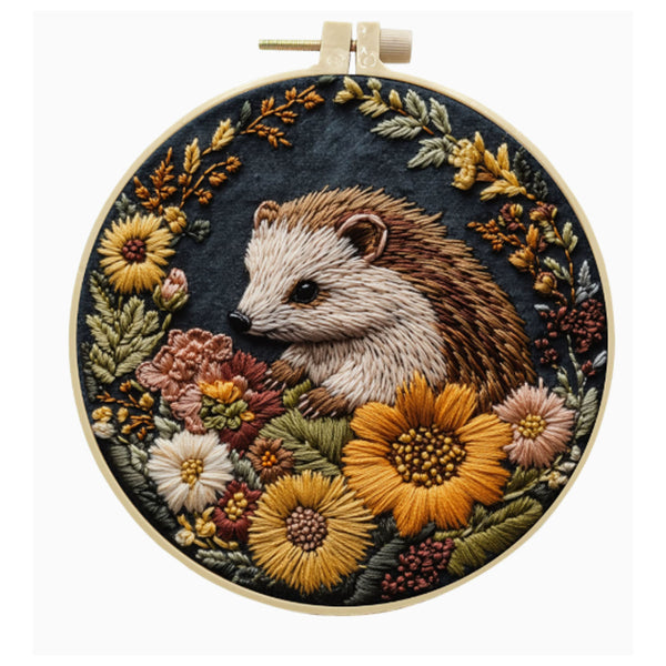 A circular embroidery hoop containing a detailed floral and hedgehog textile artwork. The design features a brown and cream hedgehog surrounded by yellow, pink, and white flowers with green and gold leaves on a dark navy background.