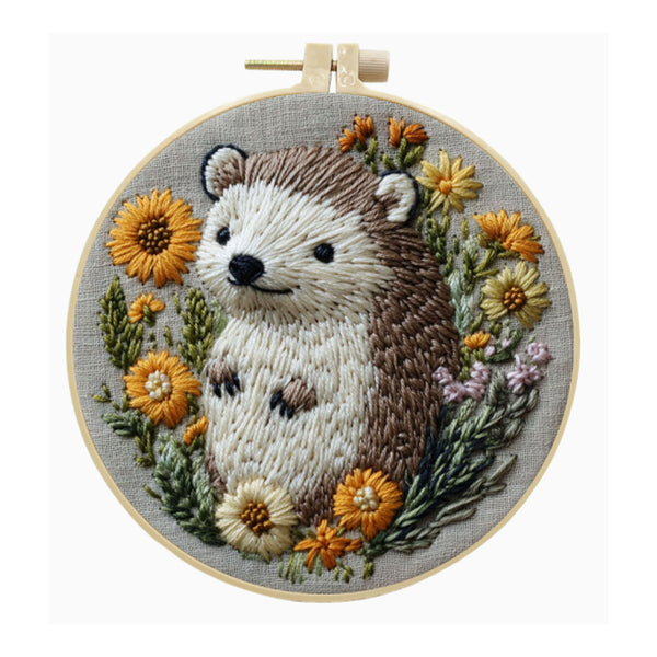 A circular embroidery hoop containing a finished embroidered hedgehog surrounded by yellow, orange, and white flowers on a light gray fabric background.