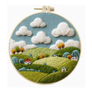 A circular embroidery hoop displaying a stylized embroidered landscape with rolling green hills, fluffy white clouds, a blue sky, and small houses and trees.