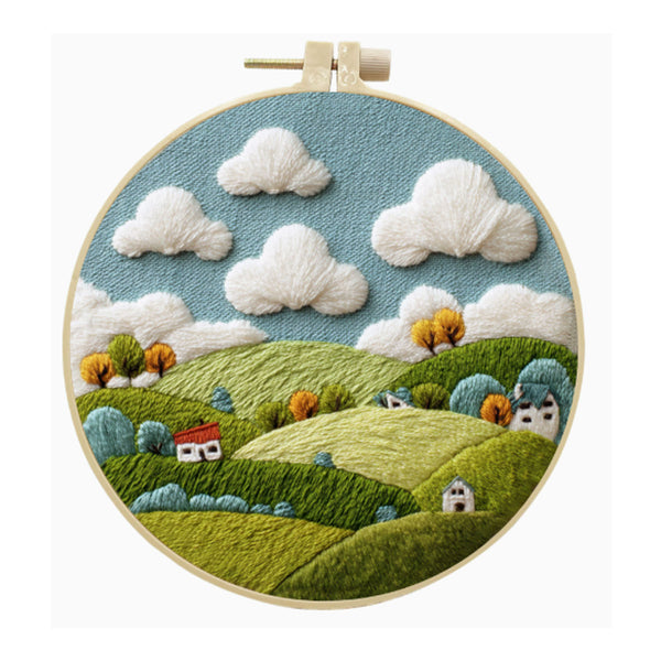 A circular embroidery hoop displaying a stylized embroidered landscape with rolling green hills, fluffy white clouds, a blue sky, and small houses and trees.