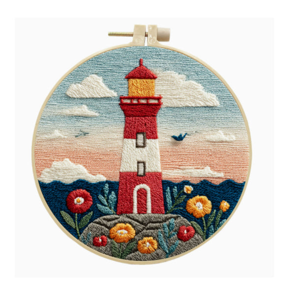A circular embroidery hoop containing a finished embroidered scene of a red and white lighthouse on a rocky shore, with blue sky, clouds, flowers, and dark blue hills. The hoop is centered on a plain white background.