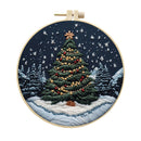 A circular embroidery hoop containing a winter scene stitched with a decorated Christmas tree, snowy ground, pine trees, and a dark blue night sky with white snowflakes.
