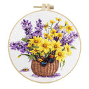 A circular embroidery hoop displaying a floral design of yellow daisies, purple lavender, a brown wicker basket, and an orange and blue butterfly on a white fabric background.