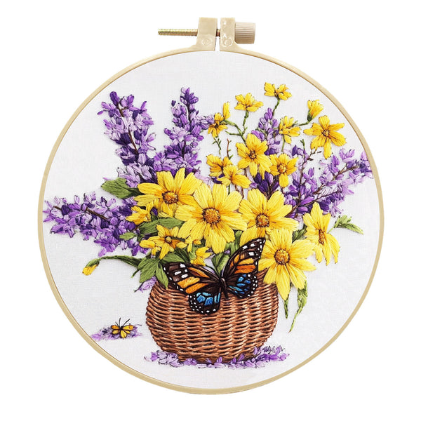 A circular embroidery hoop displaying a floral design of yellow daisies, purple lavender, a brown wicker basket, and an orange and blue butterfly on a white fabric background.