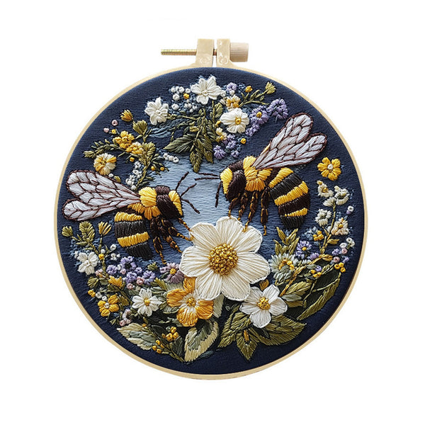 A circular embroidered needlework piece in a wooden hoop, featuring two bumblebees and white, yellow, and purple wildflowers on a dark navy blue background.