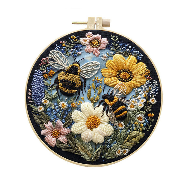 A circular embroidered floral and bee artwork displayed in a wooden embroidery hoop against a plain white background. The design features two bumblebees among large white and yellow flowers, pink blossoms, blue and purple flowers, and green leaves on a dark blue ground.