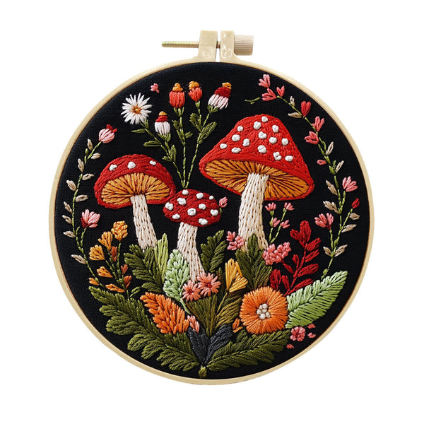 A circular embroidery hoop containing a detailed floral and mushroom embroidery design on black fabric, with a wooden hoop and white tension adjuster visible at the top, isolated on a white background.