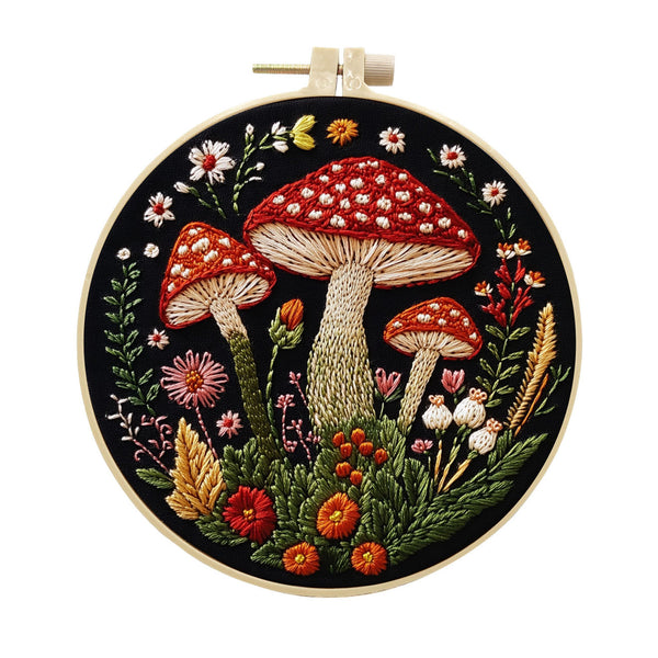 A circular embroidery hoop containing a detailed floral and mushroom embroidery design on a black fabric background. The design features three large red-capped mushrooms with white spots, surrounded by green leaves, small flowers, and wild grasses in red, orange, pink, yellow, and white.