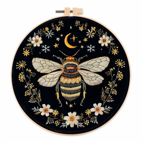 An embroidery hoop featuring a detailed stitched design of a bumblebee, a crescent moon, stars, and white flowers on a black background.