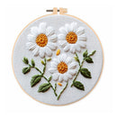 A circular embroidery hoop holding a white fabric panel with three embroidered daisy flowers, green leaves, and yellow-orange centers on a white background.