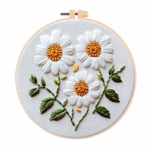 A circular embroidery hoop holding a white fabric panel with three embroidered daisy flowers, green leaves, and yellow-orange centers on a white background.
