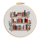 A circular embroidery hoop displaying a stitched design of a bookshelf with colorful books and potted plants on a light fabric background.