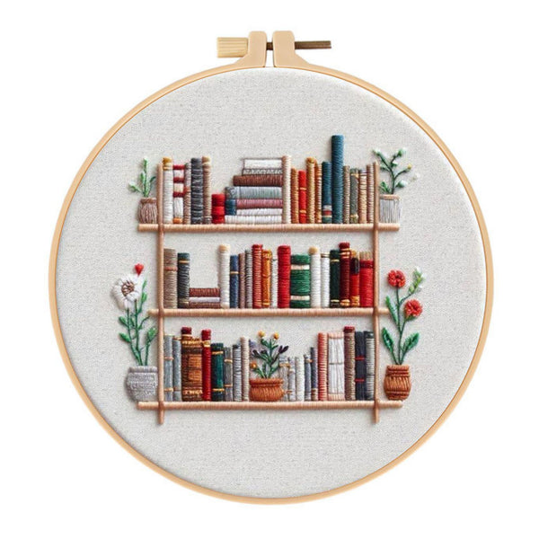 A circular embroidery hoop displaying a stitched design of a bookshelf with colorful books and potted plants on a light fabric background.