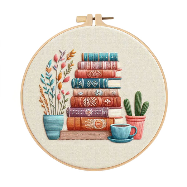 A circular embroidery hoop containing a stitched illustration of stacked vintage books, a potted plant, a cactus, and a blue cup on a light fabric background.