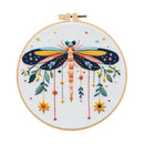 A completed embroidery piece featuring a large dragonfly with yellow, orange, and blue wings, surrounded by hanging flowers and leaves, mounted on a wooden hoop against a white background.
