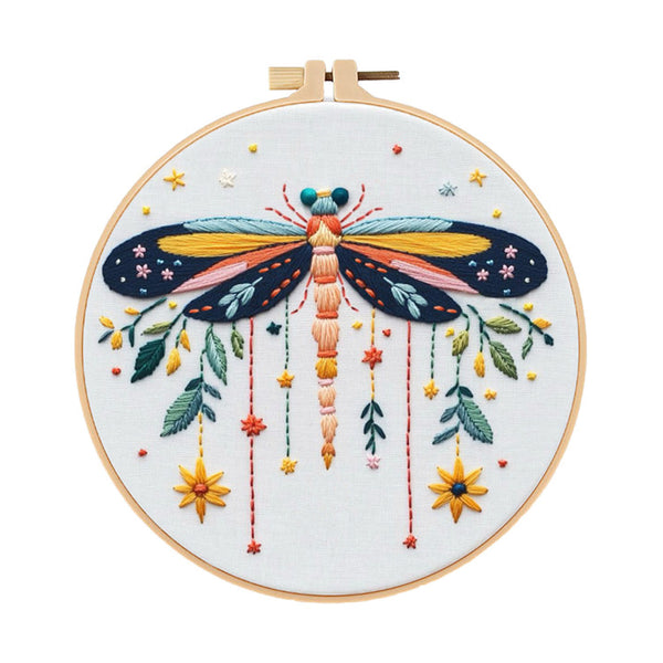 A completed embroidery piece featuring a large dragonfly with yellow, orange, and blue wings, surrounded by hanging flowers and leaves, mounted on a wooden hoop against a white background.