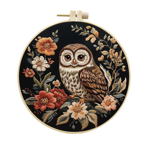 A circular embroidery hoop containing a detailed needlepoint or embroidered design of an owl surrounded by flowers on a black background, photographed against a plain white background.