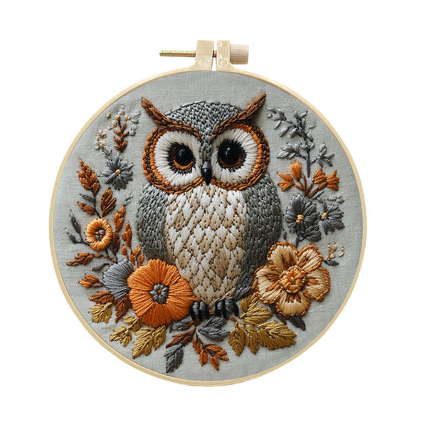 A completed embroidery piece featuring a detailed owl surrounded by autumn leaves and flowers, mounted on a light wood hoop against a plain white background.
