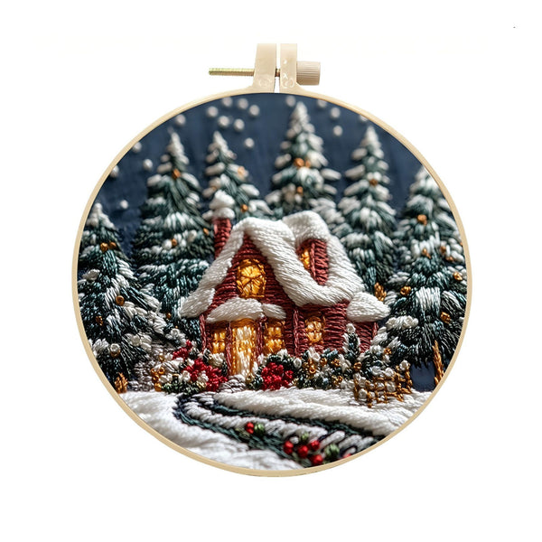 A circular embroidery hoop displaying a detailed winter village scene with a snow-covered cottage, glowing windows, pine trees, and a snowy path on a white background.