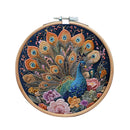 A circular wooden embroidery hoop containing a detailed peacock and floral design stitched in vibrant threads on a dark navy background, shown against a plain white background.