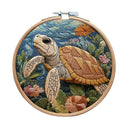 A circular wooden embroidery hoop holding a detailed embroidered sea turtle scene with blue water, green seaweed, and pink and orange flowers, shown on a plain white background.