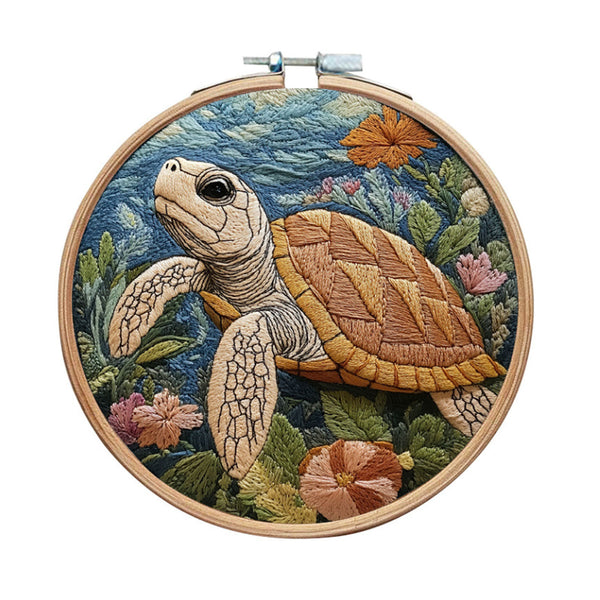 A circular wooden embroidery hoop holding a detailed embroidered sea turtle scene with blue water, green seaweed, and pink and orange flowers, shown on a plain white background.