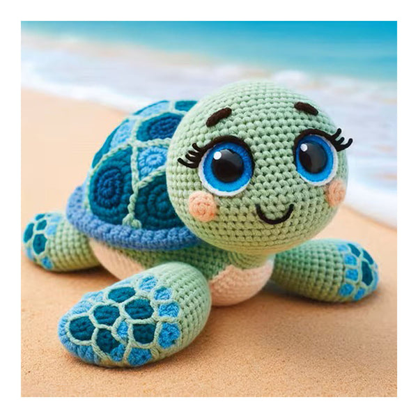 A crocheted sea turtle toy with a light green head and flippers, a blue patterned shell, and large blue eyes, posed on a sandy beach with a blurred ocean background.
