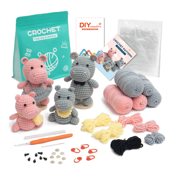 Crochet hippo toy kit with three crocheted hippo toys, a teal bag of yarn, rolled pink and gray yarn, yellow and black yarn, crochet hooks, stitch markers, clips, and pattern booklets on a white background.