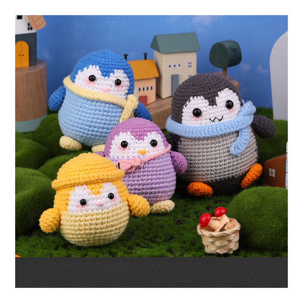 Four crocheted penguin toys in different colors and outfits, arranged on a green moss-like surface with small toy houses and trees in the background.