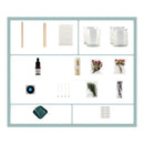A product kit arranged in a grid on a white background with a pale blue border. The kit includes two wooden spatulas, white cotton pads, two clear glass jars, a small amber dropper bottle, a jar of dried botanicals, two small flower bouquets in packets, a round blue-and-black item, four metal pins, dried green and purple botanical pieces, a dark green square item, and a sheet of white labels.