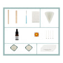 A product kit laid out on a white background with a light blue border, including wooden spatulas, a blue-handled tool, a translucent cone-shaped mold, a small amber dropper bottle, a square orange-and-yellow object, four thin metal rods, two square samples of white powder, and a card that reads THANKYOU.
