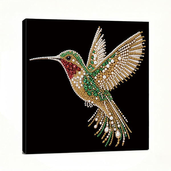 A square black canvas wall art print featuring a highly detailed, sparkling hummingbird embellished with gold, green, red, and white rhinestones, shown from a three-quarter angle against a white background.