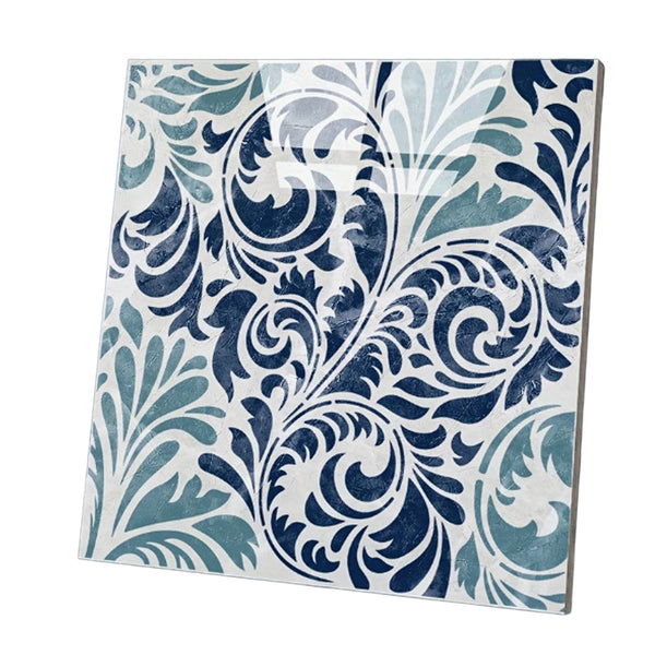 A square decorative tile featuring an ornate baroque-style scroll and leaf pattern in navy blue, teal, and white, shown at an angle on a plain white background.
