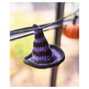 A small crocheted witch hat ornament in black and purple, hanging from a dark horizontal bar against a softly blurred outdoor background.