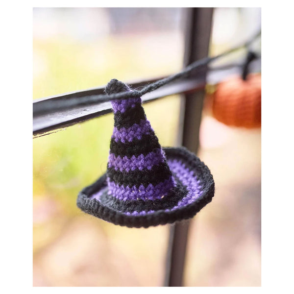 A small crocheted witch hat ornament in black and purple, hanging from a dark horizontal bar against a softly blurred outdoor background.