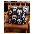 A square black and white crocheted throw pillow with a skull pattern, resting on a wooden spindle-back chair in front of a bookshelf.